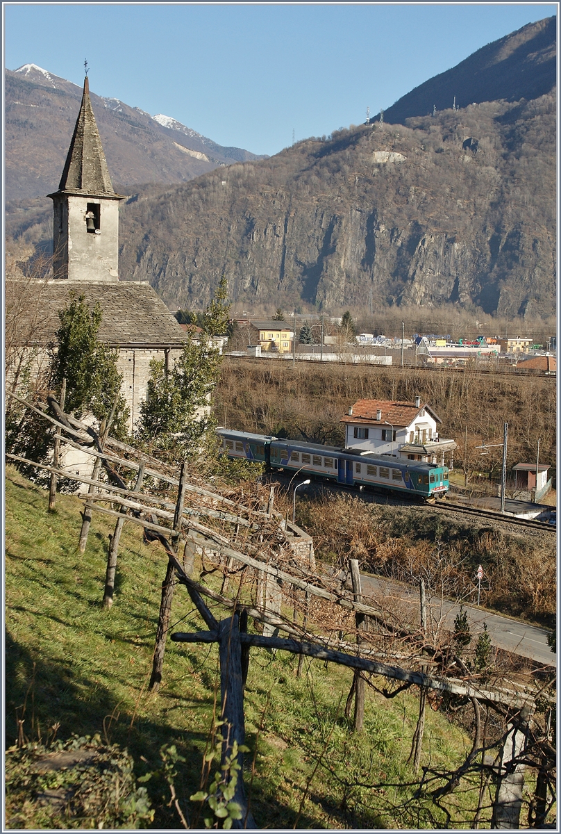 Ein aus Aln 663 Triebwagen bestehender Trenitalia Regionalzug nach Novara fährt an der aus dem 11. Jahrhundert stammenden Kirche von San Quirico vorbei. 
Das Bild entstand am südlichen Ortsrand von Domodossalo und zeigt in der Bildmitte auch noch die Hauptlinie nach Milano (RFI 23). 
17. Dez. 2013