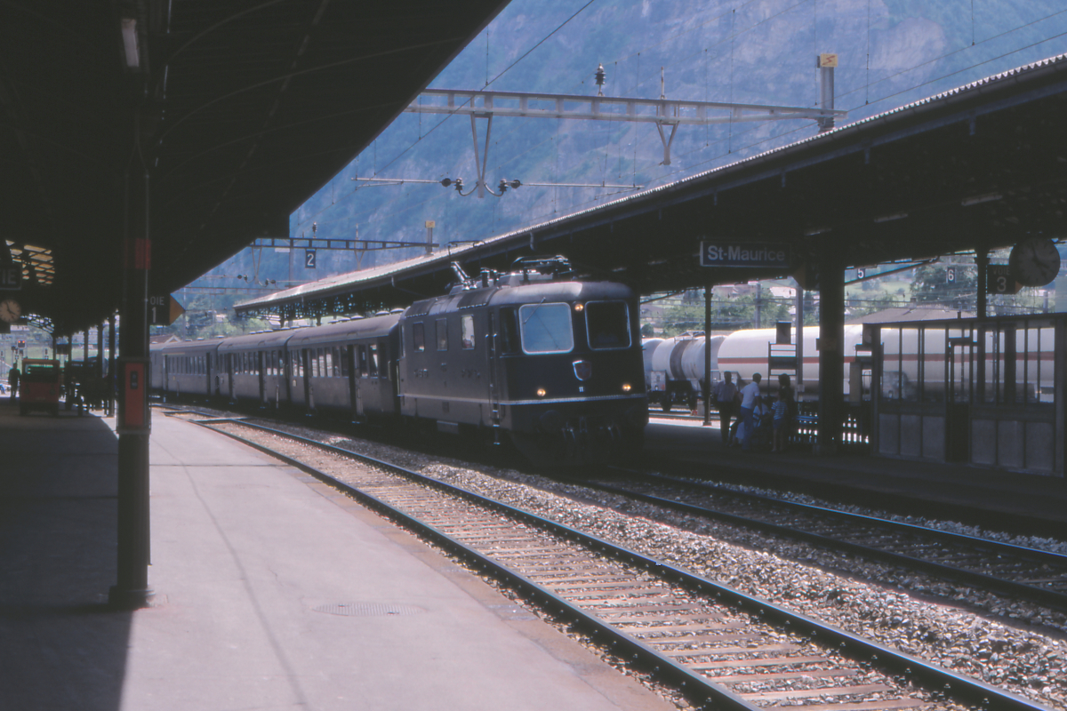 Ein Regionalzug Brig - Lausanne, geführt von Lok Re 4/4 II 11246 der SBB, fährt in Saint Maurice an der Rhone ein. Hinter der Lok laufen zwei Einheitswagen älterer Bauart, ein EW II 1.und 2. Klasse sowie ein Gepäckwagen. Der Regionalzug wartete hier fahrplanmäßig auf Überholung durch einen Schnellzug, der auch von einer Re 4/4 II geführt wurde. 
Saint Maurice, 1. Juli 1988; Canon AE-1, Canoscan, Gimp