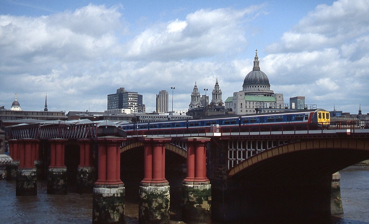 Ein Triebzug der Class 319 überquert im Sommer 1992 die Blackfriars Railway Bridge in London. Im Vorgrund die Reste der älteren, 1864 errichteten Brücke, die bis 1971 ebenfalls für den Zugverkehr genutzt und 1984 bis auf die Pfeiler abgerissen wurde, im Hintergrund die St. Pauls Cathedral.