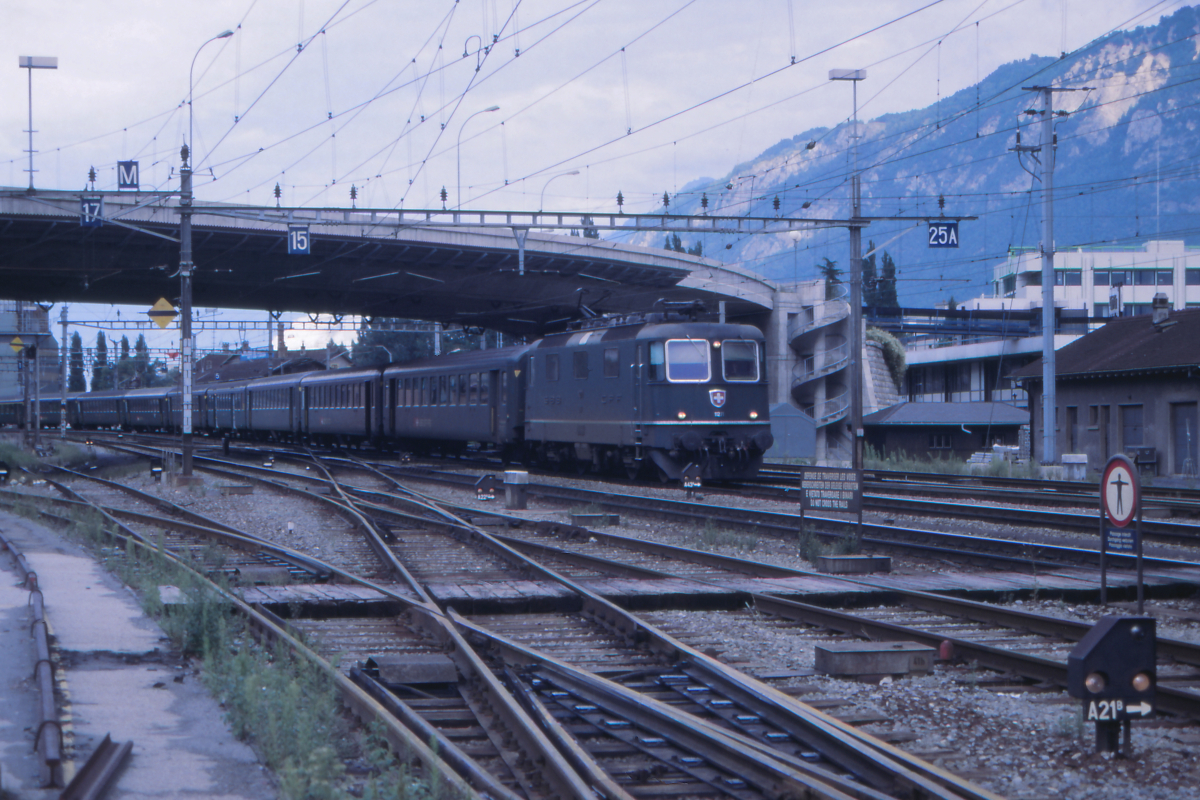 Ein typischer schweizer Schnellzug der späten 1980er Jahre: Mit einem langen Schnellzug aus Brig trifft die Re 4/4 II 11271 in Sion im Rhonetal ein. Nach einem kurzen Aufenthalt geht es weiter nach Lausanne und über Biel nach Basel oder Zürich - Romanshorn. Der Zug besteht vorwiegend aus Einheitswagen 1 (und 2), aber an vierter Stelle ist ein sogenannter Leichtstahlwagen zu erkennen, dessen Konstruktion auf die 1930er Jahre zurück geht.
Sion, 23. August 1987; Canon AE-1, Canoscan, Gimp