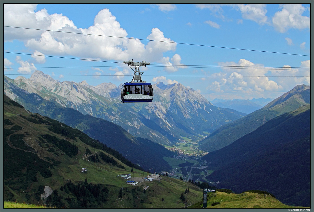 Eine Gondel der Vallugabahn bewegt sich am 09.08.2015 vor der eindrucksvollen Kulisse der Lechtaler Alpen von der Talstation auf dem Galzig bei St. Anton Richtung Valluga. Im Hintergrund (rechts der Gondel) ist die 3036 m hohe Parseierspitze zu sehen, am linken Bildrand die 2889 m hohe Vorderseespitze. Im Vordergrund ist der Gampen mit den Stationen der Mattunbahn, Kapallbahn, Gampenbahn und Nassereinbahn zu sehen. Durch das Tal schlängelt sich die 1884 eröffnete Arlbergbahn.