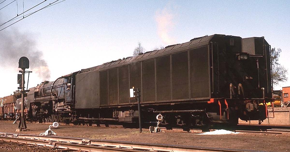 Eine Kondenslokomotive der Klasse 25 der SAR steht im November 1976 abfahrbereit in Kimberley. Bei diesen Lokomotiven wurde der Abdampf nicht durch das Blasrohr ausgestossen, sondern über eine Turbine dem Tender zugeführt, um dort zu verdunsten. Der Wasserverbrauch konnte dadurch um ca. 90 % gesenkt werden. Deshalb waren diese Lokomotiven besonders für den Einsatz in wasserarmen Gegenden geeignet. Henschel und North British bauten 1953/1954 insgesamt 90 Class 25 vor allem für den Einsatz in südafrikanischen Karoo. 1976 waren nur noch wenige Kondensloks im Einsatz, die meisten waren bereits zu 25 NC (= non condensing) umgebaut. Obwohl sie auf Kapspur (1.067 mm) verkehrten, brauchten sich diese Lokomotiven vor normalspurigen Dampflokomotiven nicht zu verstecken. Einige Daten: Gesamtachsstand 29.000 mm, Dienstgewicht Lok und Tender ca. 234 Tonnen, Achslast 18,8 Tonnen, indizierte Leistung ca. 3.400 PS, Treibraddurchmesser 1.524 mm. Aufgrund der Konstruktion erzeugte die gewaltige Lok bei der Ausfahrt kein Auspuffgeräusch, sondern setzte sich zu meinem Erstaunen eher mit einem Sirren (der Turbine) in Bewegung.