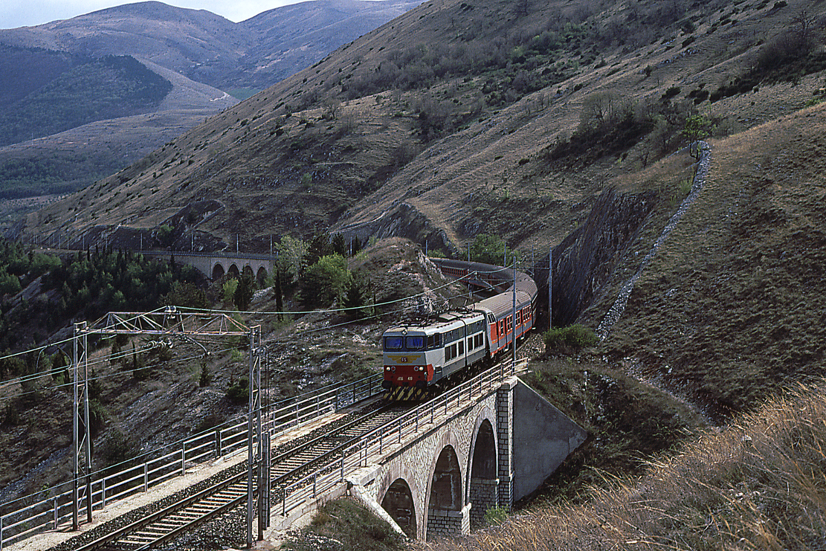 Eine der schönsten Eisenbahnstrecken führt von Rom duch die Abruzzen nach Sulmona und weiter nach Pescara. Vor der Bahnreform in Italien verkehrten hier Schnellzüge - so der Expresso Pescara - Roma, geführt von Lok E656 499. Der Zug hat die Peligna - den Talkessel von Sulmona - verlassen und fährt durch die 27 Pro-Mille-Steigung zur Galeria del Carrito, die er bald erreichen wird.
Sulmona (L'Aquila), 28. März 1990.