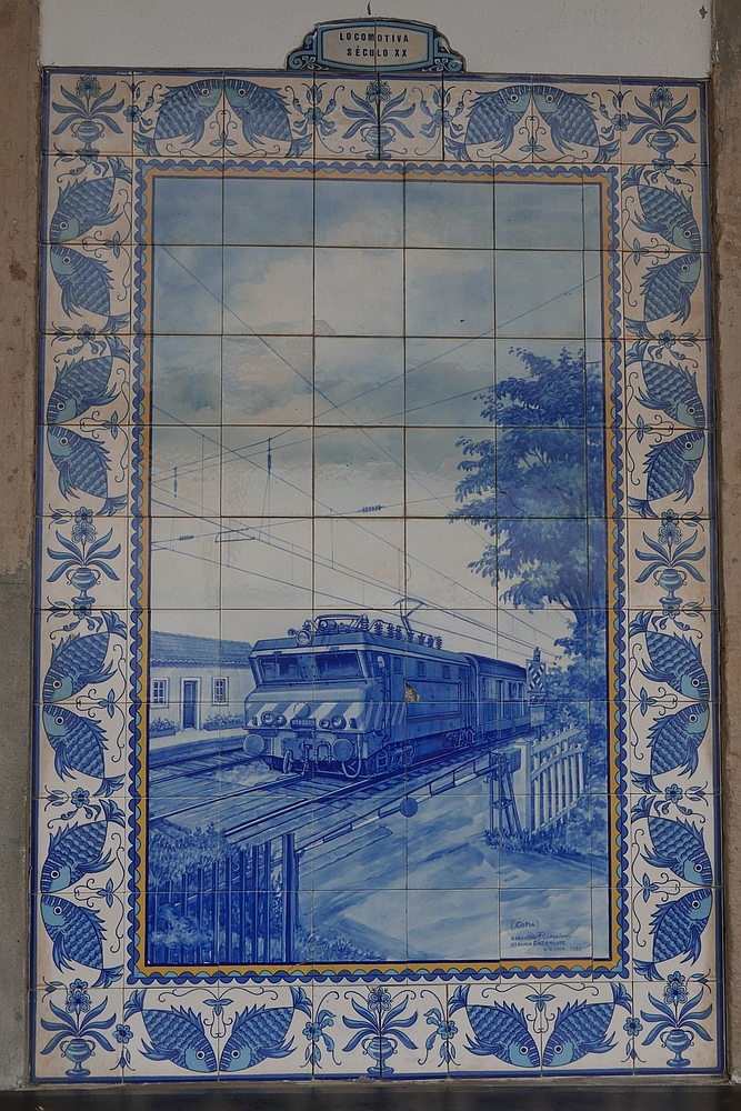 Eine Wandseite des Bahnhofgebäudes von Ovar ist mit mehreren Bildern, die aus bemalten Keramikfliesen hergestellt wurden, sogenannten Azulejos verziert. Ein Azulejo mit der Bildüberschrift „Lokomotive des 20. Jahrhunderts“ (Locomotiva século XX) zeigt eine E-Lok der Baureihe 2600 und einen Bahnübergang (11.10.2017).