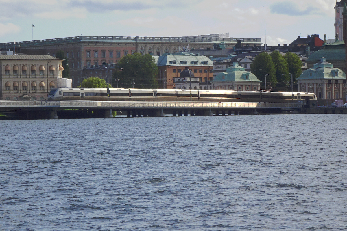 Eine wichtige Verbindung zwischen Stockholm Central und Södermalm - und weiter Göteborg und Malmö (Västra Stambanen)- bilden die Norra und Södra Järnvägsbron in Stockholm. 2021 war dort noch eine große Baustelle, 2023 sind die Eisenbahnbrücke und die parallel laufende Fußgängerbrücke fertig. Leider sind beide durch Glasscheiben getrennt.
Gerade überquert ein X55 Triebzug die Norra Järnvägsbron vor der imposanten Kulisse des Stockholmer Stadtschlosses. (Der S-Bahn-Verkehr führt in einem Tunnel unter dem Mälaren/Saltsjön hindurch.)
Stockholm, 27. August 2023