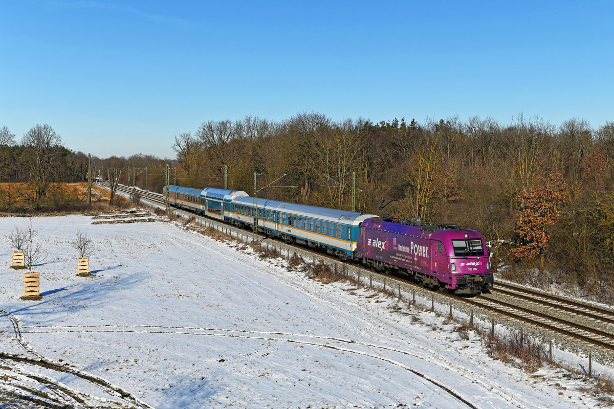 Einen kräftigen Farbakzent in der winterlich kahlen Landschaft setzte am 14. Februar 2021 die 183 005 der Länderbahn mit dem ALX 79858 von Hof nach München HBF bei Oberschleissheim. Folgenden Hintergrund hat die auffäliige Beklebung in pink und lila sowie die Umbenennung in  Alexa : Einmal feiert die Länderbahn ihr gutes Abschneiden bei einem Ranking zum Thema Gleichstellung und Diversity. Und zweitens möchte man aufgrund Personalmangels auch mehr Frauen für die Arbeit bei der Eisenbahn begeistern. 