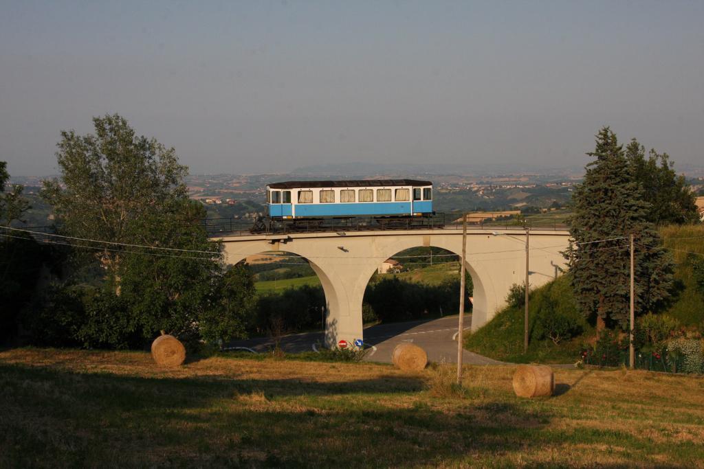 Einst führte eine elektrische Schmalspurbahn von Rimini hinauf nach San Marino. Da die Strecke im zweiten Weltkrieg stark zerstört wurde, nahm man den Betrieb nach dem Krieg nicht mehr auf. Es gibt aber noch Relikte, die an die einstige Eisenbahn in San Marino erinnern. Blickt man von der oberen Festungsseite hinunter in Richtung Rimini und Adria, so entdeckt man unterhalb des Felsmassivs dieses Eisenbahn Denkmal. Das kleinen Viadukt wurde restauriert und man stellte ein ehemaliges Schienenfahrzeug darauf. Es handelt sich um den VB 53. Die Aufnahme machte ich am Abend des 8.6.2015.