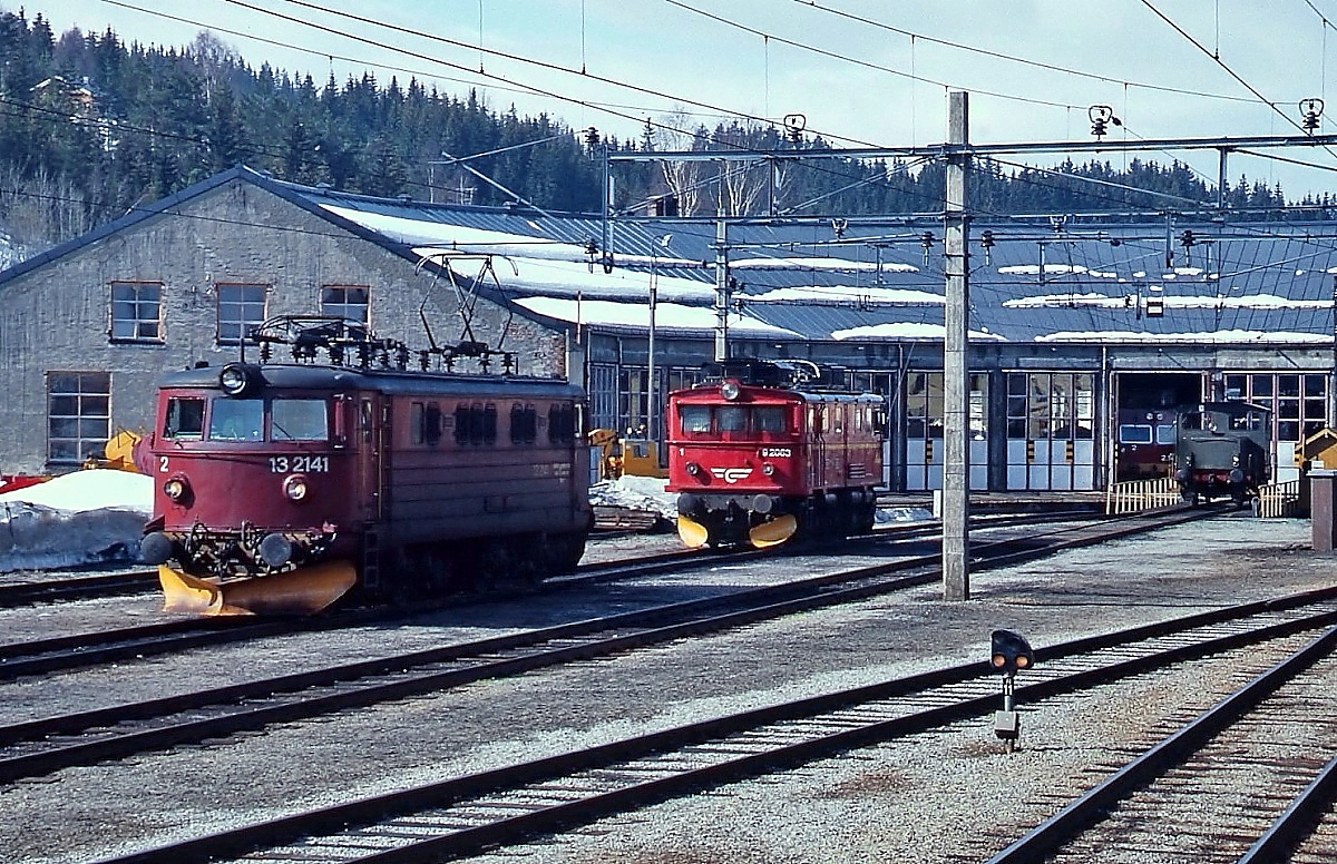 El 13.2141 und El 9.2063 Anfang Mai 1988 vor dem Lokschuppen in Geilo an der Bergenbahn, auf der Drehscheibe im Hintergrund ein Rangiertraktor der Reihe Skd 220a. Die drei Lokomotiven der Reihe 9 wurden 1944 speziell für die Flambahn gebaut und standen dort bis 1987 im Einsatz, die El 9.2063 ist heute Denkmal in Flam. El 13.2141 wurde 1961 in Dienst gestellt und 1996 abgestellt.