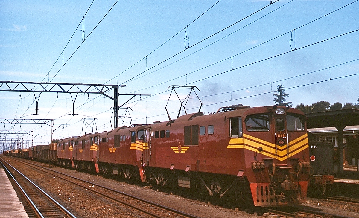 Elektrischer Betrieb in Südafrika im November 1976: Zwischen 1969 und 1985 stellte die SAR insgesamt 11 Serien der Class 6E in Dienst. Eine Dreifachtraktion mit einer weiteren abgebügelten 6E1 durchfährt hier den Bahnhof Witbank (seit 2006 eMahlaleni) an der Strecke von Pretoria nach Maputo/Mosambik, vorne die aus der 5. Serie stammende E1563 (überarbeitete Version eines bereits veröffentlichten Bildes).