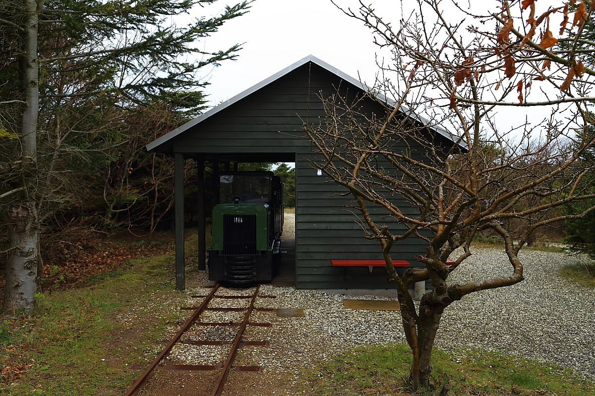 Feldbahnidylle pur! Der kleine Zug, welcher hier im  Bahnhof  der Museumsbahn Hanstholm steht, wird in wenigen Minuten, mit meinem Kollegen und mir an Bord, zu seiner ersten heutigen Fahrt starten. Die Aufnahme entstand am Vormittag des 17.04.2014, leider bei, mein übliches Pech, Regenwetter und stürmischem Wind!
Nur wenige Meter entfernt befindet sich übrigens das eigentliche Bunkermuseum Hanstholm. Hier werden in einer sehr interessanten Ausstellung viele Relikte der einstigen Festung aus der Zeit des II. Weltkrieges gezeigt und deren Geschichte erzählt. Auch gelangt man von diesem in einen original erhaltenen Geschützbunker mit Mannschaftsunterkünften, Sanitäranlagen, Funkstation, Feuerleitstand, Munitionsdepot und der riesigen Geschützanlage (Durchmesser 70 Meter!!) Nur die eigentliche Kanone fehlt. Diese kann aber am Eingang des Museumscenters besichtigt werden und ist wahrhaft gewaltig!!!