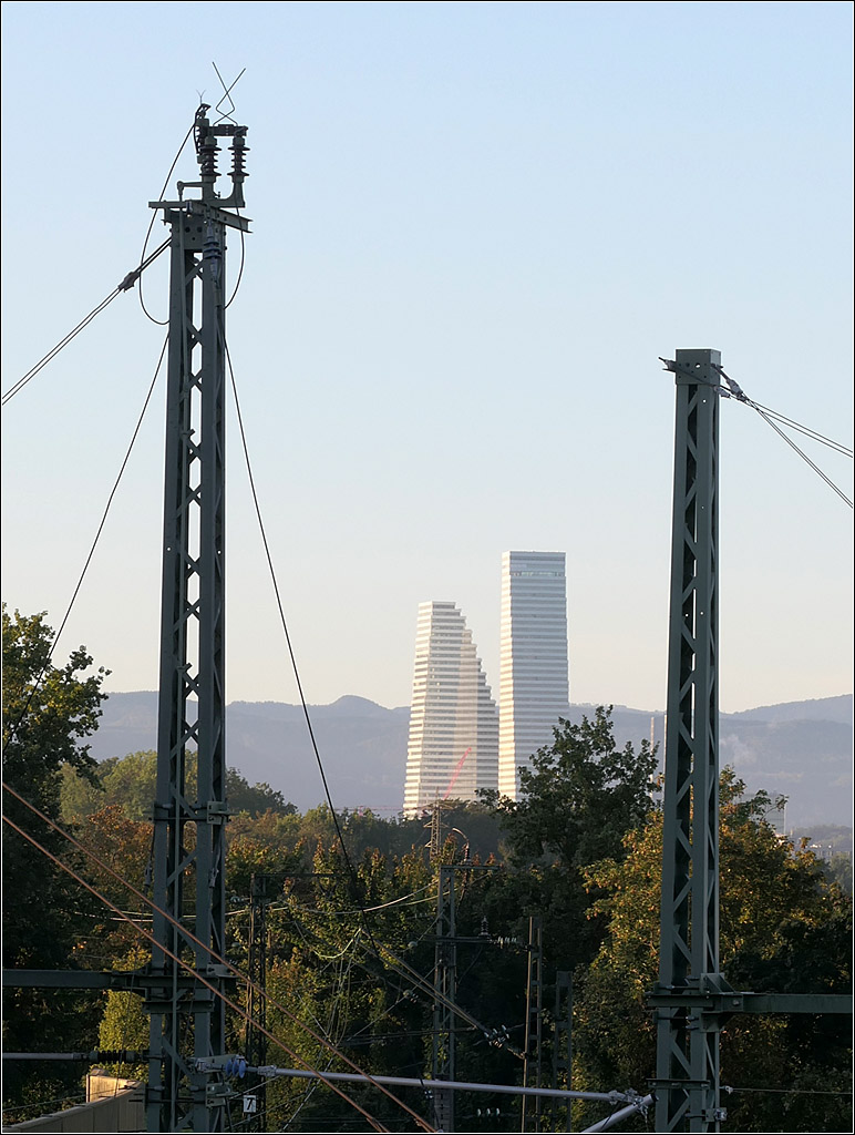 Flankiert von zwei hohen Oberleitungsmasten - 

... fällt der Blick auf die beiden Basler Roche-Türme im Licht der Morgensonne. Im Bereich der Mast zweigt die eingleisige Bahnstrecke nach Lörrach im Bahnhofsbereich in Weil am Rhein ab. Der Fotostandort befindet sich auf der Bahnbrücke der Bundesstraße 317.

18.09.2025 (M)