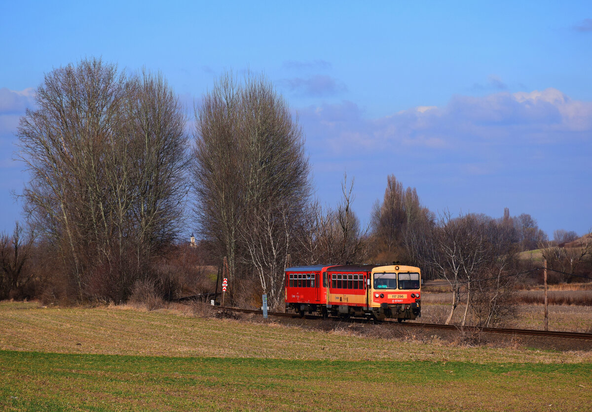 Früh-Frühling-Idyll auf der Bakonybahn: die 117 296 (Bzmot 296) als Zug 39514 von Győr nach Veszprém bei Écs.
Écs, 26.02.2022.