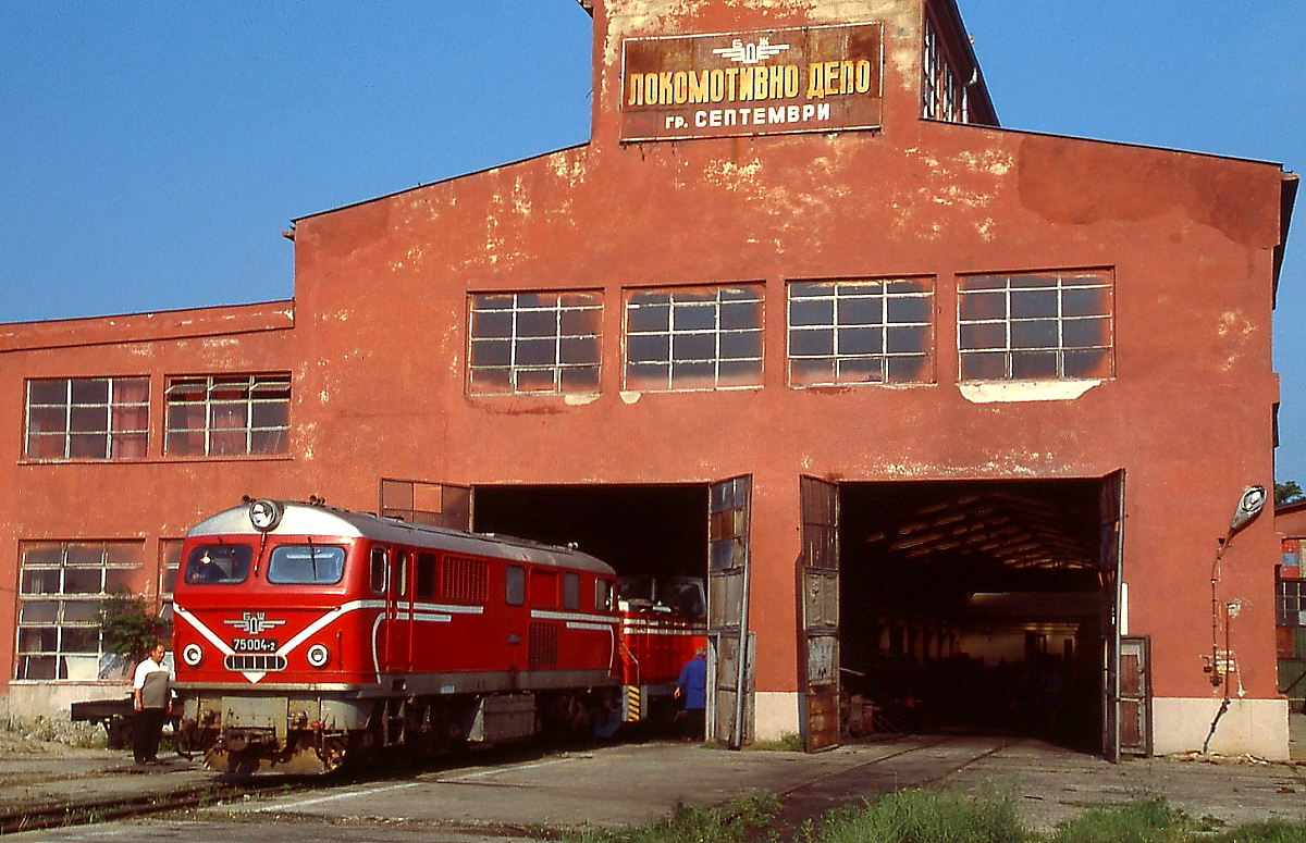 Für eine 760 mm-Schmalspurbahn hat das Lokomotivdepot Septemvri der Rhodopenbahn beachtliche Ausmaße. Am 09.06.2003 wurde hier die 1965 von Henschel gebaute 75 004-2 abgelichtet. Die Bahn verläuft zwischen Rila- und Rhodopengebirge und verbindet Septemvri an der Bahnstrecke von Sofia nach Plovdiv mit dem rund 110 km entfernte Dobrinischte. Um den Höhenunterschied von rund 1.000 m zwischen Septemvri und dem Scheitelpunkt in Awramovo zu überwinden, wurden zahlreiche Tunnels und vier Kehrschleifen angelegt.
