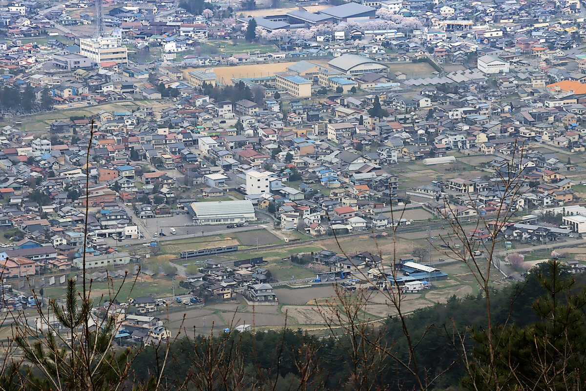 Ganz winzig - Blick auf ein japanisches Bergstädtchen. Gerade fährt ein Zug Serie 127 aus Shinano Ômachi in den japanischen Alpen aus. 21.April 2022  
