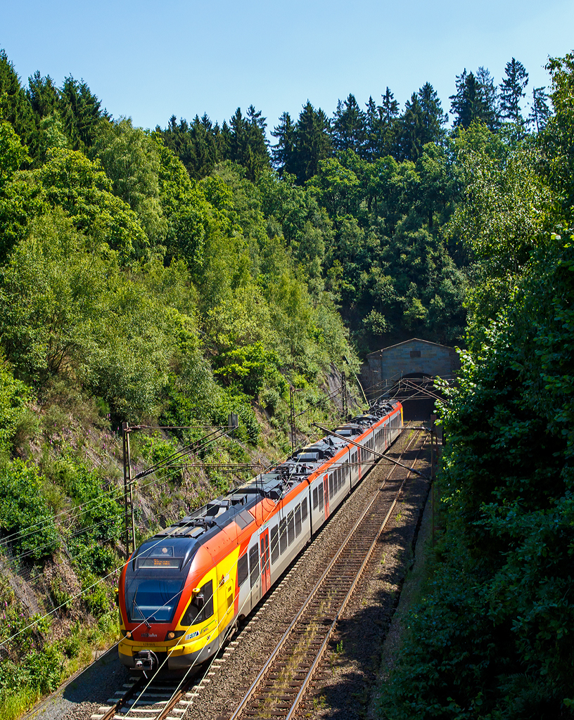
Genau 100 Jahre nach Fertigstellung kommt er nun aus dem Tunnel....
Der 5-teilige Stadler Flirt 429 045 / 545 der HLB (Hessischen Landesbahn) hat gerade (am 02.07.2015) den 2.652m langen Rudersdorfer Tunnel verlassen und fährt als RE 99 Gießen – Siegen (in Hessen, sprich auf der anderen Seite vom Tunnel. wird es als RE 40 geführt), wieder in Richtung der Endstation Siegen.

Der Rudersdorfer Tunnel ist ein 2.652 m langer Eisenbahntunnel der Dillstrecke (KBS 445) zwischen Siegen und Haiger. Als Hauptingenieurbauwerk des nördlichen Abschnitts dieser Bahnstrecke durchsticht er die Tiefenrother Höhe (552,3 m), einem Berg des Rothaargebirges auf der Grenze von Nordrhein-Westfalen und Hessen.

Aufgrund der schwierigen Topografie und der begrenzten technischen Möglichkeiten war eine direkte Verbindung zwischen Siegen, Haiger und Dillenburg zum Zeitpunkt des Baus der Deutz-Gießener Bahn vom heutigen Köln-Deutz nach Gießen in den 1850er Jahren noch nicht möglich. Erst 1915 wurde die direkte Verbindung zwischen Siegen und Haiger fertiggestellt. Am 2. Juli 1915 endeten auch die Arbeiten am Tunnel, dies sind nun genau 100 Jahre her.