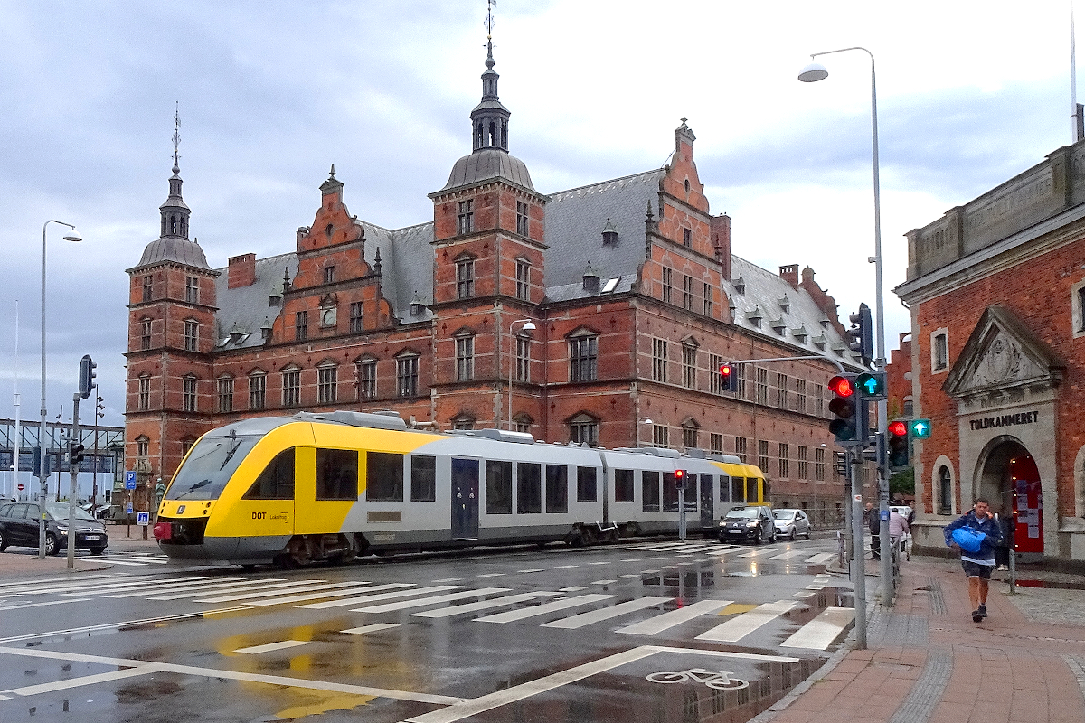 Gleich einer Straßenbahn fährt der Zug nach Gilleleje am mächtigen Hauptbahnhof von Helsingör vorbei. Vor der Inbetriebnahme der Öresundbrücke wurden von Helsingör aus Züge mit Fährschiffen über den Öresund in das nur etwas mehr als zwei Seemeilen (4 km) entfernte schwedische Helsingborg trajektiert. 27. August 2021
