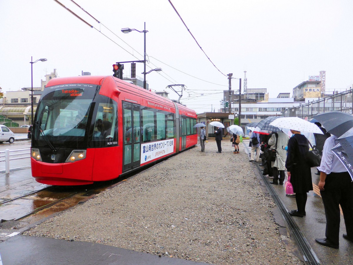 Grundangaben zur Takaoka Manyô-Linie (1067mm-Spur, Gleichstrom 600 V): Diese Linie ist in der heutigen Form als Drittsektorbahn 2001 entstanden. Dabei wurden eine Strassenbahn- und eine Ueberlandlinie zusammengelegt, nämlich die Strassenbahn von der Stadt Takaoka bis an die Küste des Japanischen Meers hinaus (8 km) und die Reste der Küstenbahn durchs Hafengebiet nach Norden (4,9 km); ursprünglich führte diese Bahn bis in die Stadt Toyama. Die Bezeichnung  Manyô  verweist auf eine berühmte Gedichtsammlung aus dem Jahre 750, in dem sich ergreifende Gedichte des damaligen Verwalters der Region um Takaoka finden und die nun an den Stationen der Bahn angebracht sind. Bild: Der Anfangspunkt vor dem Bahnhof Takaoka mit dem modernen Wagen 1001, 25.Oktober 2013. 