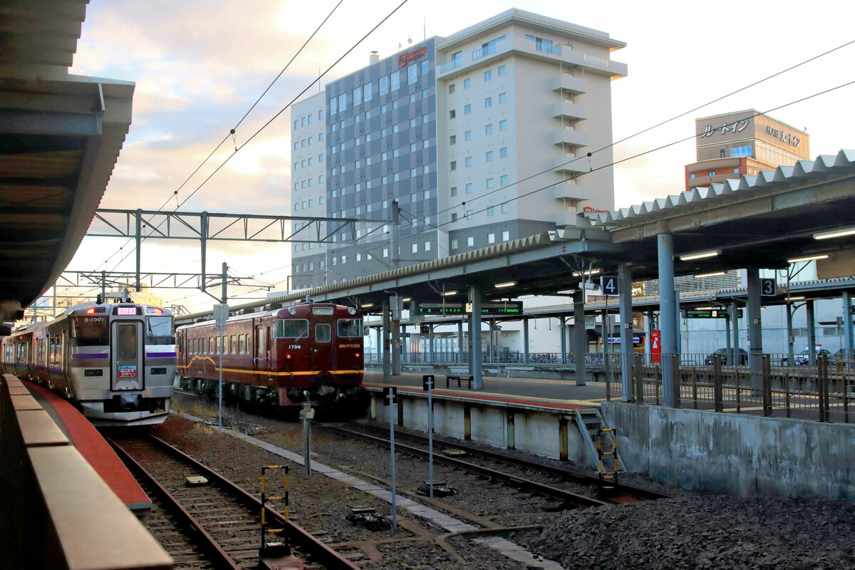 Hakodate frühmorgens im Morgenrot. Links wartet ein elektrischer Dreiwagenzug (733-1001) für die Anschlussstrecke zum Shinkansen, dessen Bahnhof etwa 18 km oberhalb der Stadt liegt. Rechts wartet ein alter Triebwagen (KIHA 40 1796) der Dônan Isaribi-Bahn, der einen Lokaldienst der Küste entlang bis Kikonai ausführen wird. 30.Oktober 2022 