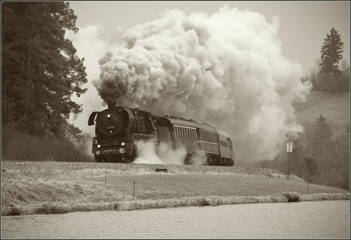 Hier kommt beim Kraftwerk Ruderatshofen die 44 546 mit viel Getöse um die Ecke. Bis Günzach werden dabei wohl noch einige Liter Dampf produziert. Februar 2018. 

Gleiches Bild wie gestern, aber ein Ausschnitt und der Schwarzweißfotografie nachempfunden. Dabei ist ein Gelbfilter, ein dem Wetter entsprechender empfindlicher aber körniger Film und ein wenig Vignette simuliert. Bei diesem Schlechtwetter Grau-in-Grau-Bild habe ich dann auch 
 gemerkt, dass die Schwarzweißfotografen eine gewaltige Palette an Einstellungsmöglichkeiten hatten, wie das Bild am Ende doch noch etwas eindrucksvoller aussah. Wenn man bedenkt, dass man das Ergebnis immer erst viel später sah, sich aber gleich auf Objektiv, Film und Filter festlegen musste, so ziehe ich nachträglich noch einmal den Hut, brauchbare Bilder waren damals durchaus mehr, als Kamera mit einigermaßen passenden Einstellungen hinhalten und abdrücken...