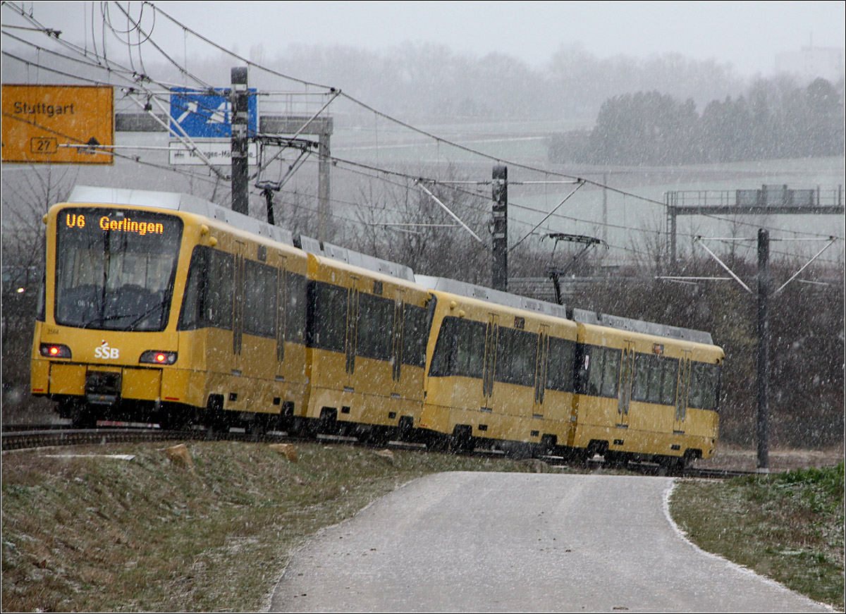 Hinab in die Talsenke -

... fährt hier ein Zug der Stuttgart U6 entsprechend dem Gefälle der natürlichen Landschaft von immerhin 60 ‰ in die Talsenke hinab. Die Strecke verläuft nahezu parallel zu autobahnähnlich ausgebauten Bundesstraße 27. Der dortige Wegweiser zeigt Stuttgart an, während das Ziel des Stadtbahnzuges mit Gerlingen angegeben wird. Bis zum Pragsattel verlaufen beide Verkehrsweg grob parallel, teilweise fährt die Stadtbahn auch direkt im Mittelstreifen dieser Bundesstraße.
Theoretisch hätte man die Stadtbahntrasse mittels Bahndamm direkt neben der B27 auf dem Neubauabschnitt führen können und sie damit völlig kreuzungsfrei anlegen können, so aber hat sie den Reiz des mehrfachen Auf und Ab und es waren weniger Erdbewegungen und Kunstbauten nötig.

07.01.2022 (M)