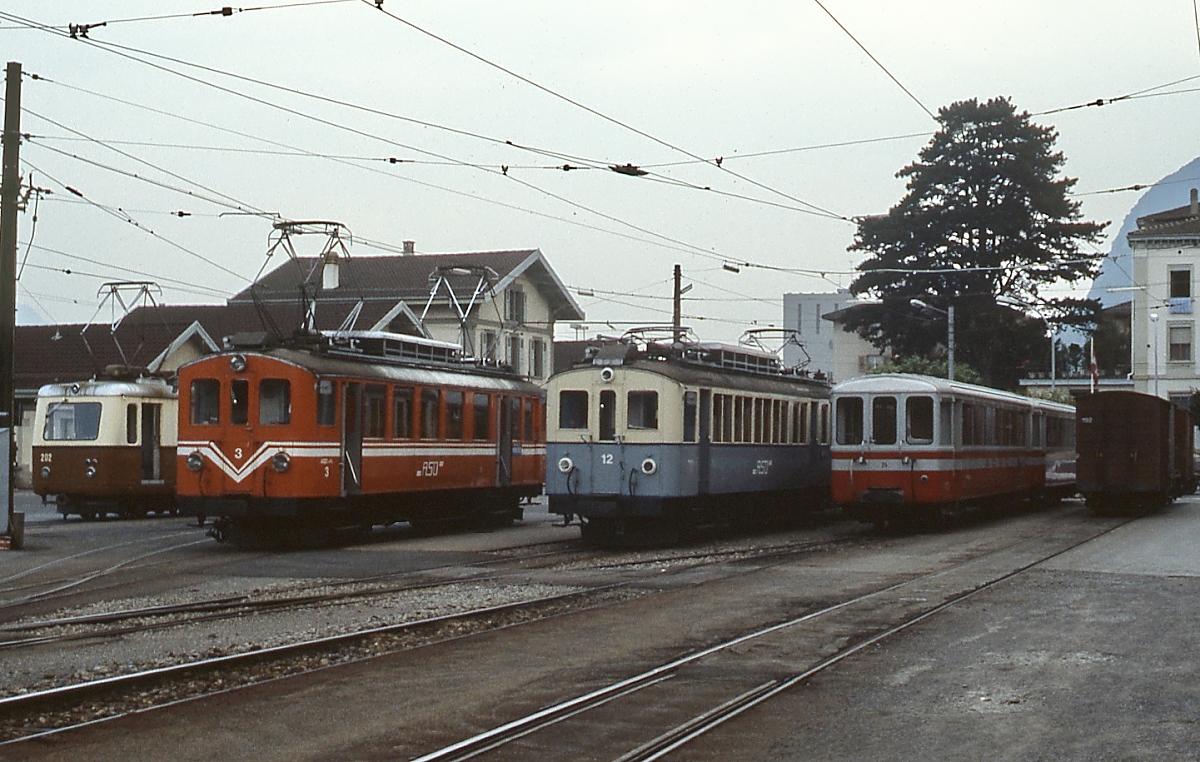 Hochbetrieb auf dem Bahnhofsvorplatz von Aigle: An einem Maiabend 1980 stehen dort (von links nach rechts) der ABDeh 2/4 202 der AL (Aigle-Leysin), daneben die ABDe 4/4  3 und 12 der ASD (Aigle-Sepey-Les Diablerets) und zwei Beiwagen der AOMC (Aigle-Ollon-Monthey-Chambery), ganz rechts noch zwei Güterwagen. Die beiden Fahrzeuge der ASD unterscheiden sich nicht nur farblich, der ABDe 4/4 3 erhielt im Rahmen einer Modernisierung auch eine andere Fensterteilung. Ebenso wie die NyStC wurde die ASD zu diesem Zeitpunkt noch mit Triebwagen aus der Zeit der Eröffnung (1913) betrieben und war - nachdem die Straße nach Les Diablerets wintersicher ausgebaut worden war - stark einstellungsgefährdet. Aber auch diese Bahn verkehrt mit modernen Fahrzeugen noch heute.