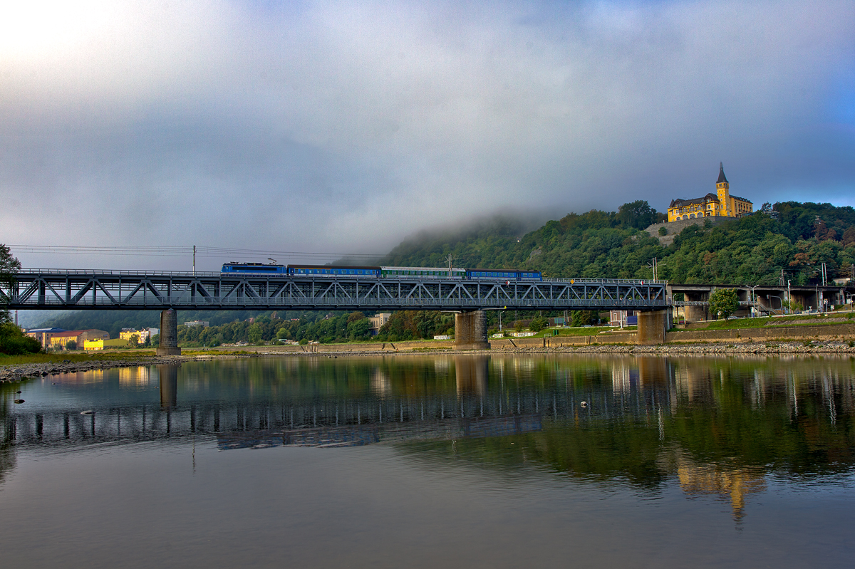 Im Elbtal setzt sich am frühen Morgen langsam die Sonne durch und die Nebelschwaden lichten sich und geben den Blick frei auf die Eisenbahnbrücke und das 4 Sterne Hotel Vĕtruše,dass über der Stadt Ústí nad Labem thront.Es ist aus der Stadt bequem mit der hauseigenen Seilbahn erreichbar,am rechten Bildrand ist die Kabine kurz vor der  Bergstation  zu erkennen.Derweil fährt die 163 077-1 mit dem R 785 über die Brücke.Bild vom 10.9.2015