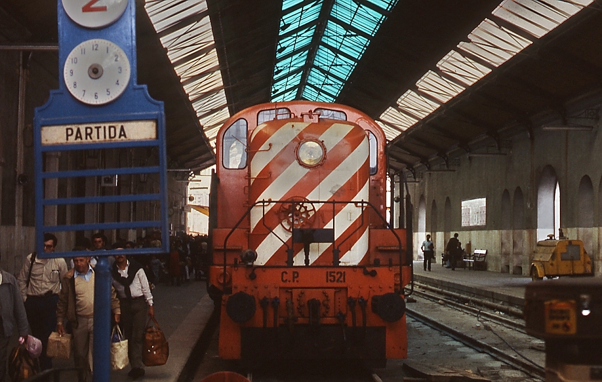 Im etwas düster wirkenden Bahnhof Barreiro ist die CP 1521 im April 1984 mit einem Personenzug angekommen. Hier endeten früher alle aus dem Süden nach Lissabon verkehrenden Züge. Seit 1999 fahren die Fernzüge über die Brücke des 25. April direkt nach Lissabon, nach dem Bau eines neuen Bahnhofs in Barreiro steht die Halle zwar noch, doch die Gleise wurden entfernt. Die 1521 gehört zu den 1948/51 von ALCo gelieferten Lokomotiven der Serie 1501-12/1521-25, die den  Road switchern  des Typs RSC 3/RSC 4 entsprechen.