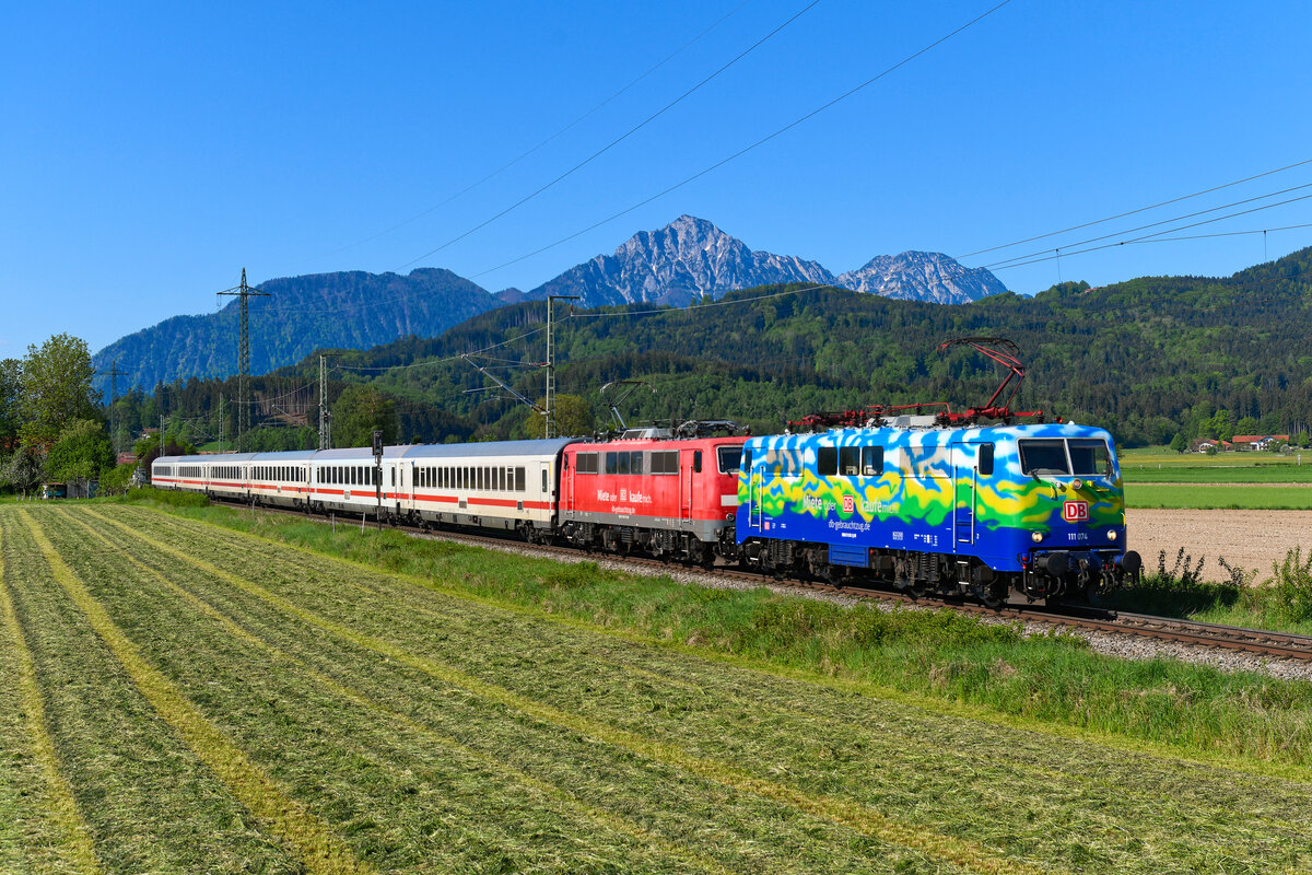 Im Jahr 2025 fanden die letzten Fahrten des Zugpaars IC 2082/83 von und nach Berchtesgaden mit Lokbespannung und klassischem Wagenpark statt. Da zuletzt die Streckenklasse herab gesetzt wurde und die Baureihe 101 somit die Zulassung auf dem Streckenabschnitt ab Freilassing verlor, behalf sich DB Fernverkehr mit der Anmietung von Loks der Baureihe 111 bei DB Gebrauchtzug. Zur Freude vieler Eisenbahnfreunde wurden dabei insbesondere die bunt gestalteten Exemplare aus dem Bestand verwendet. Unter anderem die in den Farben des ehemaligen Touristik-Zuges gehaltene 111 074. Am 01. Mai 2025 beförderte die gemeinsam mit der verkehrsroten Schwester 111 067 den IC 2082 bis Freilassing. Bei Ainring konnte ich den Zug kurz vor seinem Etappenziel mit dem Staufen-Massiv im Hintergrund und frisch gemähter Wiese im Vordergrund fotografieren. 