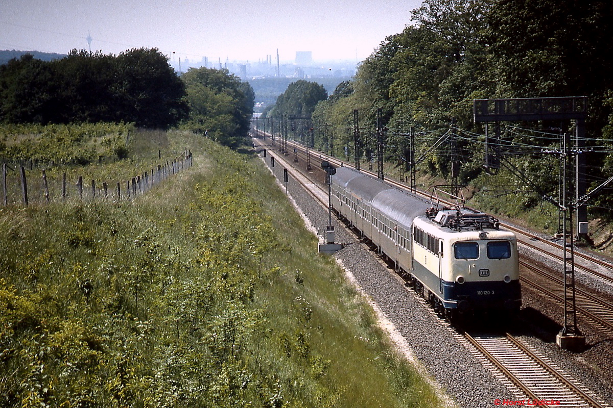 Im Juni 1987 fährt 110 120-3 die Steigung von Erkrath nach Hochdahl hinauf. Gleichzeitig auch ein (leicht verspäteter) Gruß zum 175. Geburtstag: Am 20.12.1838 eröffnete die Düsseldorf-Elberfelder Eisenbahn-Gesellschaft die Strecke von Düsseldorf nach Erkrath, 1841 wurde die Verlängerung bis Hochdahl und weiter nach Elberfeld in Betrieb genommen. Vom Bahnhof Erkrath bis Hochdahl hat die Bahn auf ca. 2,5 km 82 Höhenmeter zu überwinden. Wegen der geringen Leistungsfähigkeit der damaligen Lokomotiven wurden die Züge zunächst mittels einer stationären Dampfmaschine in Hochdahl an einem Seil nach oben gezogen. Ziemlich schnell wurde die Dampfmaschine durch eine Umlenkrolle ersetzt und talwärts fahrende Lokomotiven oder Züge unterstützten die bergwärts fahrenden mittels Seil. Erst 1926 wurde dieses System aufgegeben, bis zur Elektrifizierung wurden bis zu drei Dampflokomotiven bei schweren Zügen für die Bergfahrt benötigt.