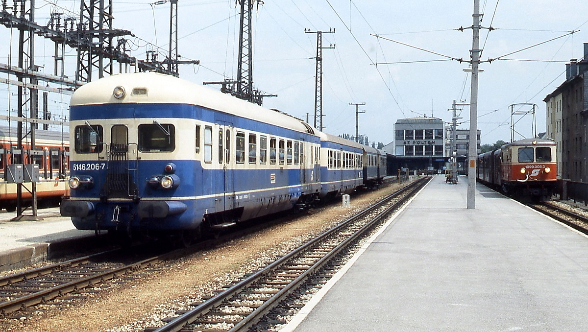 Im Juni 1987 steht 5146.206-7 abfahrtbereit vor einem Regionalzug in St. Pölten Hauptbahnhof. Hinter dem Steuerwagen sind noch zwei in Triebwagenfarben lackierte Zweiachser angehängt. Rechts steht 1099.008-3 vor einem Personenzug der Mariazellerbahn.
