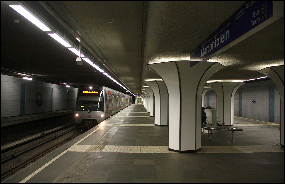 Im Untergrund von Rotterdam -

Die dreigleisige Station Marconiplein an der hauptsächlich in Ost-West-Richtung durch Rotterdam verlaufenden Calandlijn. Der erste Abschnitt dieser zweiten Rotterdamer Metrolinie wurde 1982 eröffnet. 1983/84 wurde die Linie in Richtung Nordosten erweitert, aber als Stadtbahn (Sneltram) in Niveaulage mit Kreuzungen durch Auto- und Fußgängerverkehr. Auf diesem Sneltram-Abschnitt erfolgt die Stromzufuhr über Oberleitungen und nicht über Stromschienen wie im 'echten' Metrobereich.

Die unterirdische Verlängerung nach Westen bis zu der Station Marconiplein ging 1986 in Betrieb. Hier kann zu einigen Straßenbahnlinien umgestiegen werden. Die Station hat drei Gleise, wobei das dritte Gleis nur einen Hilfsbahnsteig hat, links auf dem Bild erkennbar. Gestalterisch wirksam sind die später eingebauten pilzförmigen Verkleidungen der Stützen mit indirekter Beleuchtunggen an der Decke. 

2002 wurde ab hier die Strecke über Schiedam und das Hafengebiet bis Tussenwater verlängert, von wo die Calandlijn gemeinsam mit der Erasmuslijn bis Spijkenisse de Akkers verkehrt.

21.06.2016 (M)