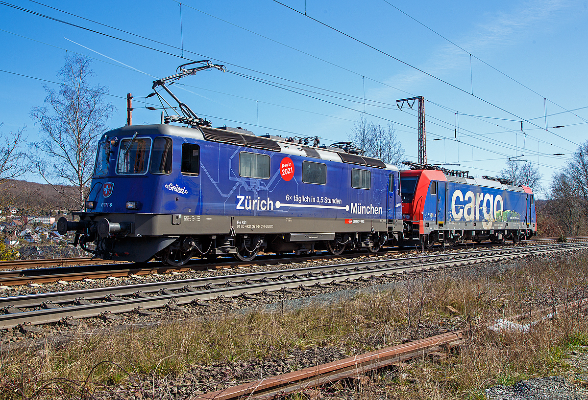 Ja, „Grüezi“ oder „Servus“.....
Die SBB Cargo Re 421 371-6 (91 85 4421 371-6 CH-SBBC), ex Re 4/4 II 11371, mit Werbung Zürich – München 6 x täglich in 3,5 Stunden, fährt am 30.03.2021 mit der SBB Cargo Re 484 016-1(91 85 4484 016-1 CH-SBBC), ex E 484.016 SR, einer TRAXX F140 MS am Haken durch Rudersdorf (Kr. Siegen), an der Dillstrecke (KBS 445), in Richtung Siegen.

Eine SBB Cargo Re 484 wie die 016-1 kann man eigentlich Deutschland nur mit viel Glück sehen, denn sie haben nur die Zulassungen für die Schweiz und Italien. Sie haben keinen Stromabnehmer mit breiterer Wippe gemäß deutscher Vorschrift, auch die die deutsche Zugsicherung Indusi fehlt ihnen. Der Lz war auf dem Weg nach Krefeld.

Lebensläufe:
Die Re 421 371-6 wurde 1983 von der SLM Winterthur (Elektrik BBC/ SAAS) unter der Fabriknummer 5235 gebaut und als Re 4/4 II 11371 an die SBB geliefert. Im Jahr 2002 erfolgte der Umbau zur Re 421 für die SBB Cargo (Einsatz auch in Deutschland). Die zugelassene Höchstgeschwindigkeit für die Schweiz beträgt 140 km/h und für Deutschland 120 km/h.

Die Re 484 016-1 eine Bombardier TRAXX F140 MS wurde 2004 von Bombardier in Kassel unter der Fabriknummer 34009 gebaut und an die SBB Cargo AG, als E 484.016 SR geliefert. 