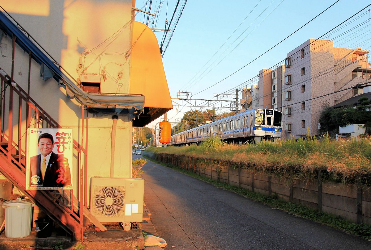 Japanische Stadtquartiere - Zug 1755 des Odakyû-Konzerns in Ikuta in der Agglomeration Tokyo. Ein freundlicher Mann auf Stimmenfang an einem Restaurant, das den Geist schon aufgegeben hat - im Abendlicht des 18.Oktober 2015. 