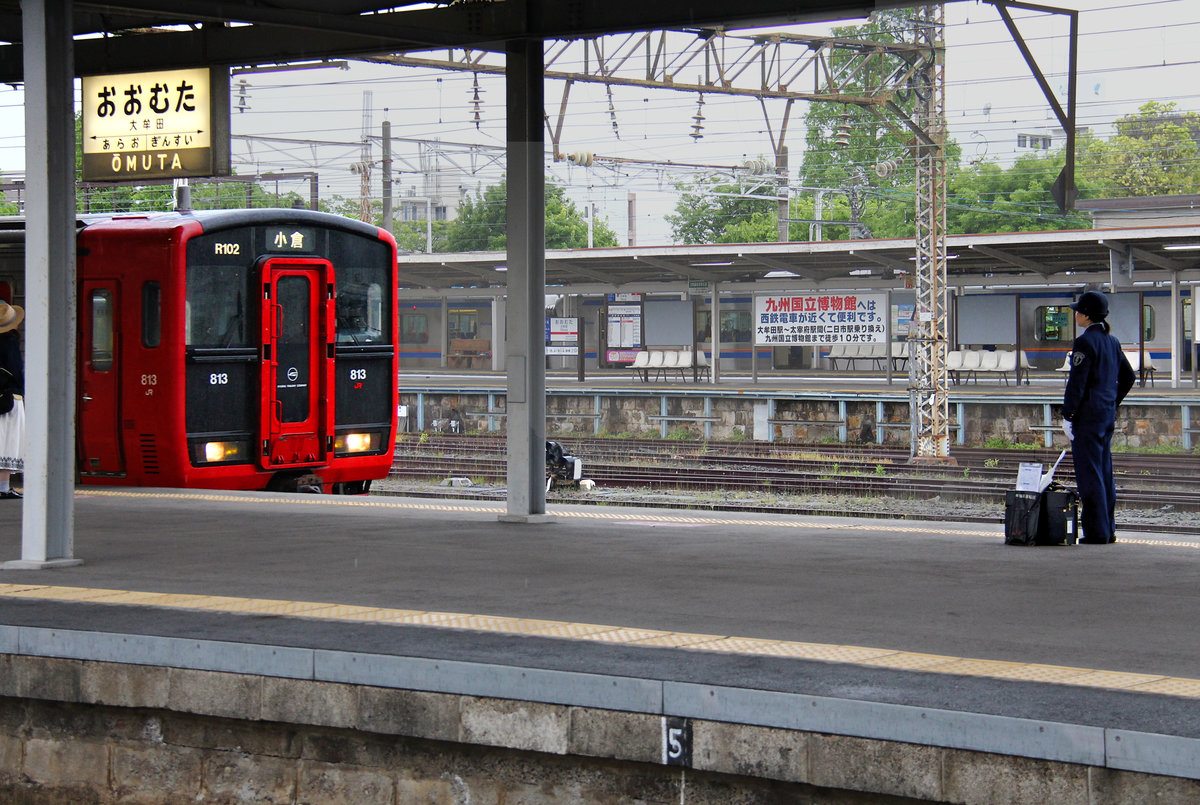 JR Kyûshû Serie 813: Die Wagenführerin, die Zug 813-102 für die Fahrt nach Fukuoka übernimmt, wartet am genau vorgeschriebenen Platz in der Station Ômuta an der Nordwestküste von Kyûshû. Sie kontrolliert dabei die Stirnfront des einfahrenden Zugs. Zu ihrer Ausrüstung gehört neben dem grossen Dienstfahrplan (den sie während der Fahrt mit dem Finger und mit lautem Ausrufen verfolgen wird) auch ein weisser Schirm. 3.Mai 2016  