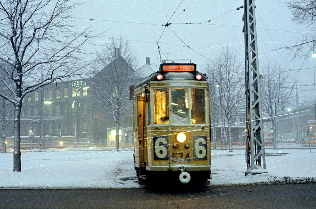 København / Kopenhagen Københavns Sporveje SL 6 (Tw 574 + Bw 15xx) Østerbro (København Ø), Jagtvej / Lyngbyvej / Vibenhus Runddel am 26. Februar 1969. - Trotz des geritzten Negativs habe ich beschlossen, dieses Bild hochzuladen, um ein Foto zu zeigen, das nur durch eine Zusammenarbeit mit freundlichen Personen zustande gekommen ist. Ein Freund, der auch Straßenbahnfreund war und ein Auto zu Verfügung hatte, hatte vorgeschlagen, dass wir herumfahren könnten, um einige Straßenbahnen im schönen Schneewetter zu machen. Mit dem Wagenführer des fotografierten Zuges hatten wir dann verabredet, er solle einige Sekunden am Vibenhus Runddel, einem großen Platz, stillhalten, damit wir ein Abendfoto (auf etwa zehn Sekunden mit Stativ und Drahtauslöser) machen könnten. Die Nachmittags-HVZ war noch nicht vorüber, und es gab noch viel Autoverkehr, weshalb wir damit würden rechnen können, große Probleme während des Fotografierens zu bekommen. Zu der Zeit wurde der Verkehr am Ort noch von Polizeibeamten reguliert, und dann haben wir einen Beamten gefragt, ob er die Autos zurückhalten wollte, um uns die Möglichkeit zu geben, ein Bild zu machen - er hat ja gesagt und uns dadurch geholfen. Das Bild ist also nur eine Realität geworden, weil mir drei hilfsbereite Personen Hilfe geleistet haben. - Wir Bahnfotografen machen viele Fotos allein, aber ab und zu gelingt dem einen oder dem anderen vielleicht etwas mehr, wenn man mit anderen zusammenarbeiten kann. - Mit diesen Worten samt diesem und dem folgenden Foto möchte ich Freunden und allen anderen Benutzern der Bahnbilder.de einen guten Rutsch ins Neue Jahr und alles Gute im Neuen Jahr wünschen. 