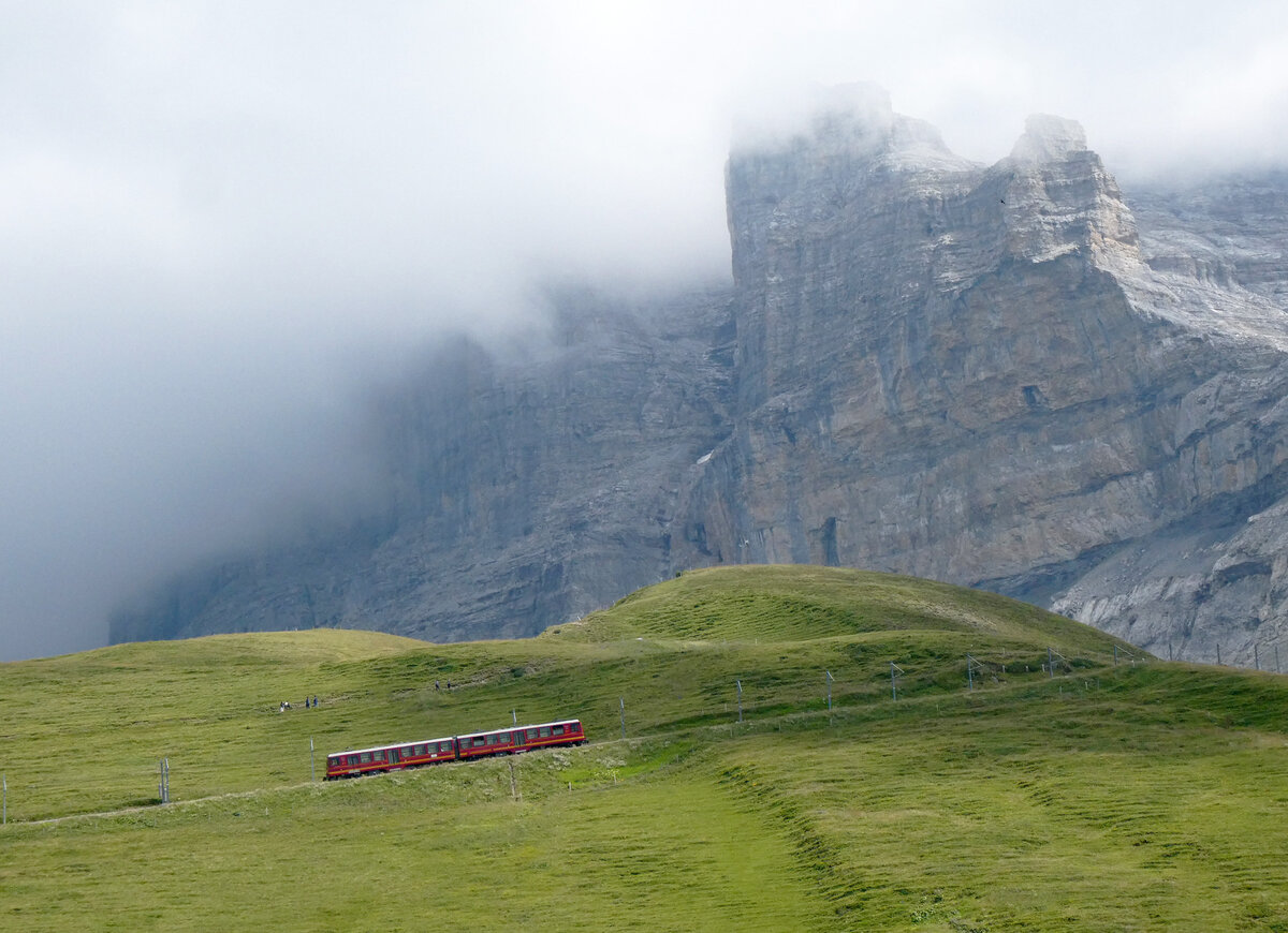 Kleiner roter Zug auf gr�ner Wiese vor grauer Felswand: Zug der JB hat die Ausgangsstation Kleine Scheidegg vor wenigen Minuten verlassen und f�hrt bergw�rts in Richtung Station Eigergletscher - Jungfraujoch. Im Hintergrund ein Teil der weltber�hmten Eigernordwand. Kleine Scheidegg, 21.7.2026