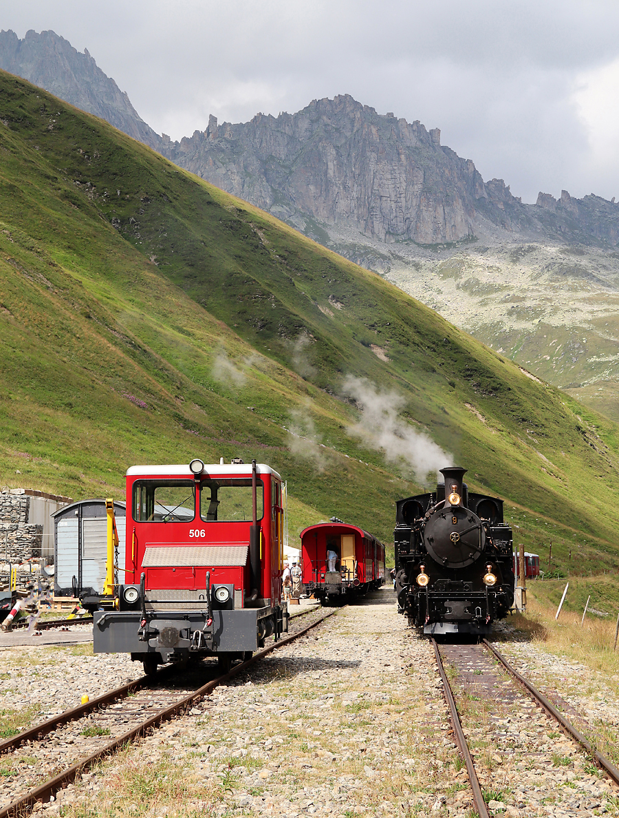 Kreuzung der beiden Dampfzüge zwischen Realp und Oberwald in der Station Furka (2160 müM). Rechts wartet die Lok BFD 9 (ex Brig-Furka-Disentis-Bahn) auf die Weiterfahrt Richtung Gletsch - Oberwald. Die Draisine 506 hat beim roten Zug im Scheiteltunnel zwischen Muttbach-Belvédère und Furka Schiebedienst geleistet. Der Scheiteltunnel ist 1.8 km lang und die Steigung beträgt in der erwähnten Fahrtrichtung während den ersten 1500 m 30 Promille. Es ist keine Zahnstange vorhanden. So kann übermässige Dampfentwicklung verhindert werden und im Notfall wäre eine Hilfslok bereits am Zugschluss. Das Foto wurde vom Publikumsbereich aus gemacht. Furka, 28.7.2022