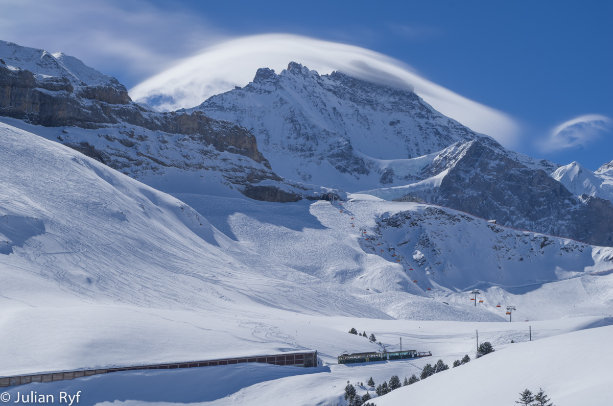 Kurz vor Mittag sind die bergwärts fahrenden Skifahrer schon nicht mehr so zahlreich. So ist der Skiwagen dieser alten Komposition geschoben von einem BDhe 4/4 der ersten Serie am 28.03.2015 nur schwach belegt und die Fahrgäste sind wohl grösstenteils Touristen auf dem Weg auf das Jungfraujoch. In Kürze erreicht der Zug die Dienststation Salzegg nur wenig unterhalb der Kleinen Scheidegg. Im Hintergrund zu sehen die Sesselbahn Eigernordwand mit den auffallenden orangen Windschutzhauben und über dem ganzen die Jungfrau, deren Gipfel von Schneeverwehungen eingehüllt ist. Gut zu erkennen auch die seit 2011 stillgelegte Richtstrahlstation Jungfraujoch auf dem Ostgrat der Jungfrau. Diese soll in Zukunft zu einem öffentlich zugänglichen Aussichtspunkt umgebaut werden.