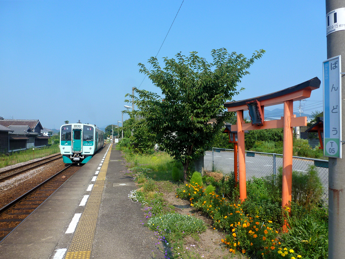 Lokalverkehr auf Shikoku - der Nordosten: Triebwagen Serie 1500 Nr. 1562 in Bandô. Auf dem Bahnsteig ein kleiner Torbogen, der den Eingang in den heiligen Bereich eines Schreins bildet (dieser befindet sich etwas weiter oben am Berghang). 10.August 2013.  