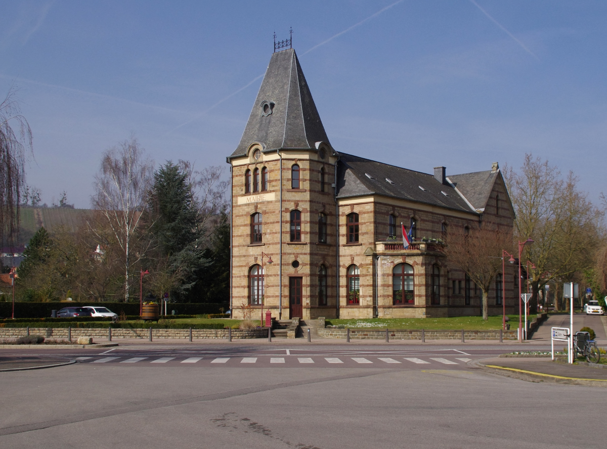 Mairie in Wasserbillig - Ein Gebäude mit Vergangenheit - Mairie steht dran und ist auch drin aber.......Wer sich den Turm des Gebäudes anschaut und mit vielen gleichartigen Gebäuden im Großraum Luxemburg - Lothringen vergleicht, der kommt auf die Lösung. Dieses Gebäude war mal ein Bahnhof. Wasserbillig gönnte sich den Luxus im Unkreis vom 300 Metern zwei Bahnhöfe zu besitzen. Das Großherzogtum Luxemburg gehörte von 1815 bis 1866 als souveräner Staat zum Deutschen Bund . Daher wurde der Bau verschiedener Eisenbahnlinien mit  deutschem  Geld bezahlt. Die Wilhelm-Luxemburg-Bahn wurde 1861 eröffnet. Diese führte von Luxemurg über Wasserbillig in Richtung Trier. Hier erfüllte ein eher nüchterner Zweckbau die Rolle des Bahnhofs.
Dieser Bahnhof nannte sich Wasserbillig GL (Guillaume-Luxembourg). Eine weitere Bahngesellschaft erreichte Wasserbillig im Jahr 1874, die Prinz-Heinrich-Eisenbahngesellschaft. Diese Strecke (Sauertalbahn) führte von Ettelbrück bis an die Mosel. die in Wasserbillig erreicht wurde und weiter bis Grevenmacher führte.
Die Prinz Heinrich Eisenbahngesellschaft baute ihren eigenen Bahnhof in unmittelbarer Nähe des anderen Bahnhofs von Wasserbillig. Dieses stattliche Gebäude trug den Namen Wasserbillig  PH  (Prinz Heinrich). Nach Stilllegung der Sauertalbahn war der Bahnhof Wasserbillig PH ohne Verwendung und wurde von der Stadtverwaltung zum Rathaus umgestaltet. Auf den Resten der Sauertalbahn findet noch auf 10 Km Zugverkehr statt. Personenverkehr zwischen Ettelbrück und Diekirch und Güterverkehr zwischen Wasserbillig und dem Moselhafen Mertert. 07.03.2014