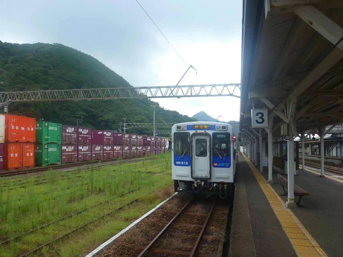 Matsuura-Bahn, Imari-Arita-Linie: Wagen 616 wartet im morgendlichen Gegenlicht im Endbahnhof Arita. Die vielen Container links stehen im  Arita Off-rail-station  und warten nicht etwa auf den Bahntransport (Güterverkehr gibt es hier längst nicht mehr), sondern auf Strassentransport. Rechts im Bild die JR-Strecke nach Haiki und Sasebo. 7.August 2013. 