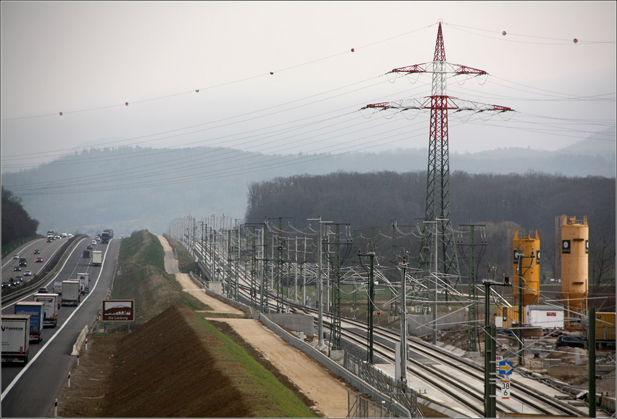 Meist an der Autobahn entlang -  

führt die Neubaustrecke Wendlingen - Ulm. Ähnlich wie die Autobahn folgen auch die Gleise dem Auf und Ab der hügeligen Landschaft. Aus dieser Perspektive wird die Strecke fast in einen Käfig eingesperrt durch die dichte Folge der Oberleitungsmasten. 
Blick auf die Strecke in Richtung Ulm nach dem östlichen Tunnelportals des Albvorlandtunnels bei Kirchheim unter Teck. 

16.03.2022 (M)

