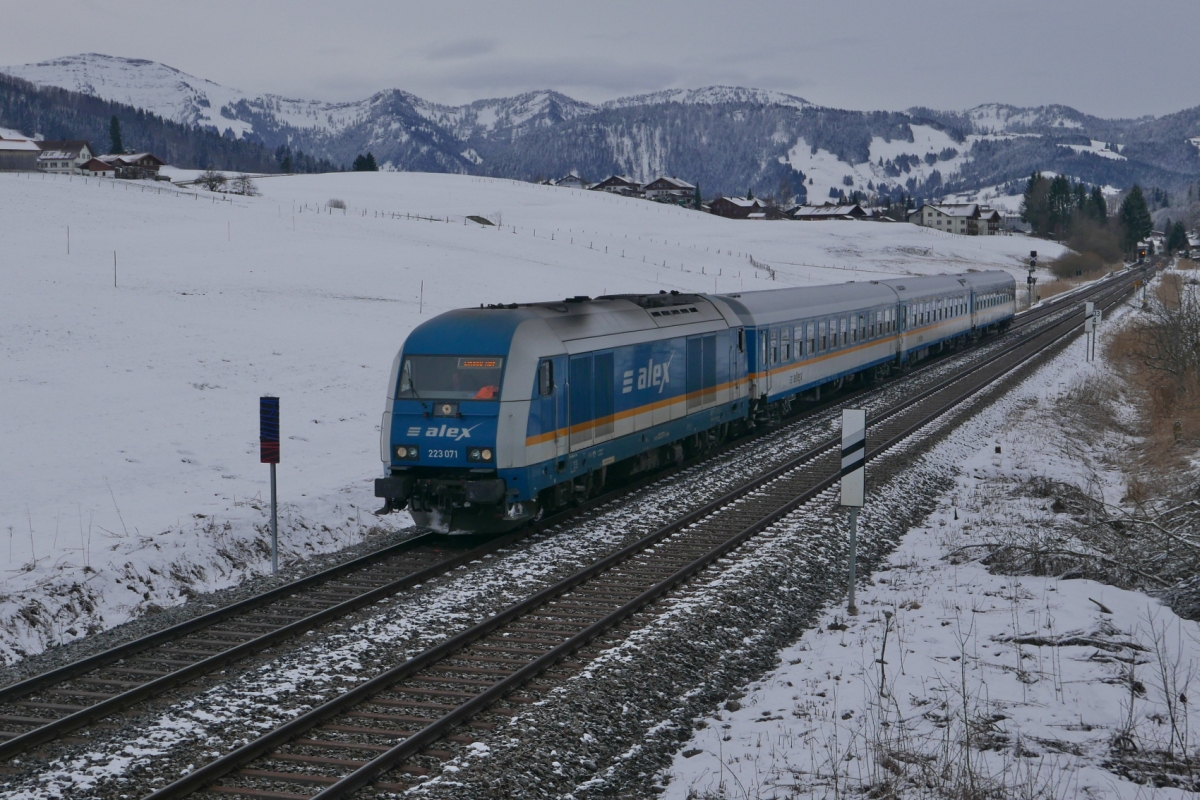 Mit Blick auf die Nagelfluhkette im Hintergrund zieht 223 071 den ALX 84132, München - Lindau, am 20.02.2016 bei Oberstaufen durch die leicht verschneite Allgäuer Landschaft.