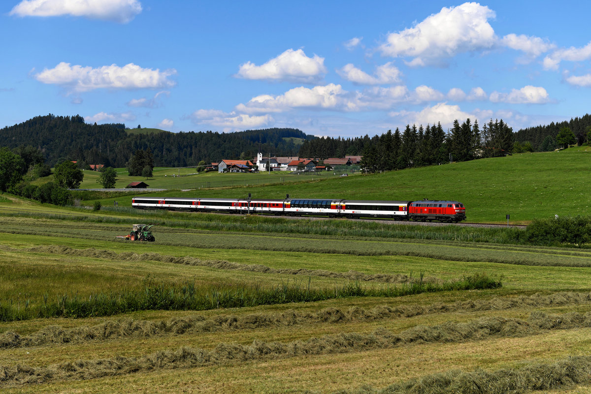 Mit sechs Wagen und nur einer Lok wies der EC 195 genau die richtige Länge für das Motiv mit Blick auf die kleine Ortschaft Zell auf. In flotter Fahrt nähert sich die 218 430 Oberstaufen und überholt dabei einen Traktor, der mit der Heuwende beschäftigt ist (25. Juni 2020).  