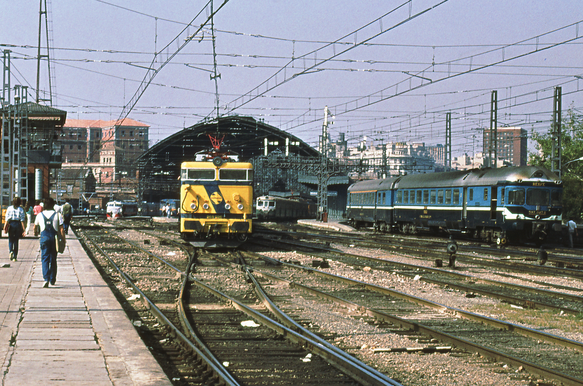 Mittags, etwa ab 13.30 Uhr, herrschte Hochbetrieb am Bahnhof Madrid Atocha. Es fuhren Züge ab in die großen andalusischen Städte und in die Extremadura. Lok 269 226, eine Vertreterin der letzten Bauserie 269 222 bis 269 331, beginnt gerade ihre Reise ins 727 km entfernte Cadiz. Am Zughaken hat sie den Talgo Pendular 110. Ganz rechts sehen wir einen TER, einen Dieseltriebwagen italienischer Herkunft, links im Hintergrund steht eine von Krauss Maffei gebaute Diesellok mit dem Talgo III nach Granada und Almeria. Oktober 1983. 
Canon AE1, Canoscan, GIMP.
