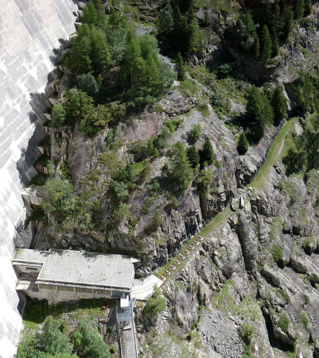 Nahaufnahme eines alten Gebäudes mit ein paar Meter Gleis, aufgenommen von der Staumauer des Lac d'Emosson aus. Das teilweise verschüttete Trassee führt ohne Gleis nach rechts in Richtung heutigem Endbahnhof des Panoramic Train Pied du Barrage. Das Foto wurde von der Staumauer aus gemacht. Lac d'Emosson, 25.8.2025