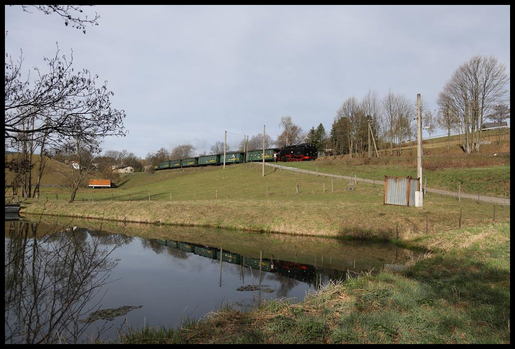 Nahe Hammerunterwiesenthal ist hier am 26.4.2019 um 8.29 Uhr die 991782-5 mit dem ersten Personenzug des Tages von Oberwiesenthal nach Cranzahl unterwegs. Im Wasser eines Fischteiches spiegelt sich der Zug im Wasser.