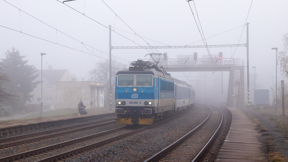 Nebeliges zum 16. November 2018 in Zelenice nad Bilinou. Der Schnellzug mit 362 068-9 durchfährt den Bahnhof auf dem Mittelgleis. 