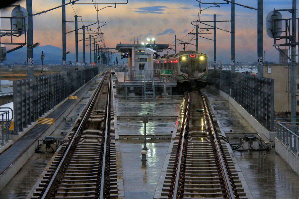 Neues Leben im Katastrophengebiet südlich von Sendai. Im Tsunami vom 11.3.2011 wurden die Bahnlinie hier, ebenso wie die Siedlungen in Meeresnähe, vollständig zertrümmert; zwei Züge der Serie E721 wurden weggeschwemmt und ebenfalls zertrümmert. Nun ist die Strecke wieder aufgebaut (Wiedereröffnung  10.Dezember 2016), jetzt aber viel weiter weg vom Meer und - wie zu sehen - über weite Strecken in Hochlage. An der ganz neuen Kreuzungsstation Yamashita warten zwei Triebzüge Serie E721. (Leider hat ein heftiger Regenguss Tropfen an der Scheibe hinterlassen, die hingenommen werden müssen). 14.November 2017. 