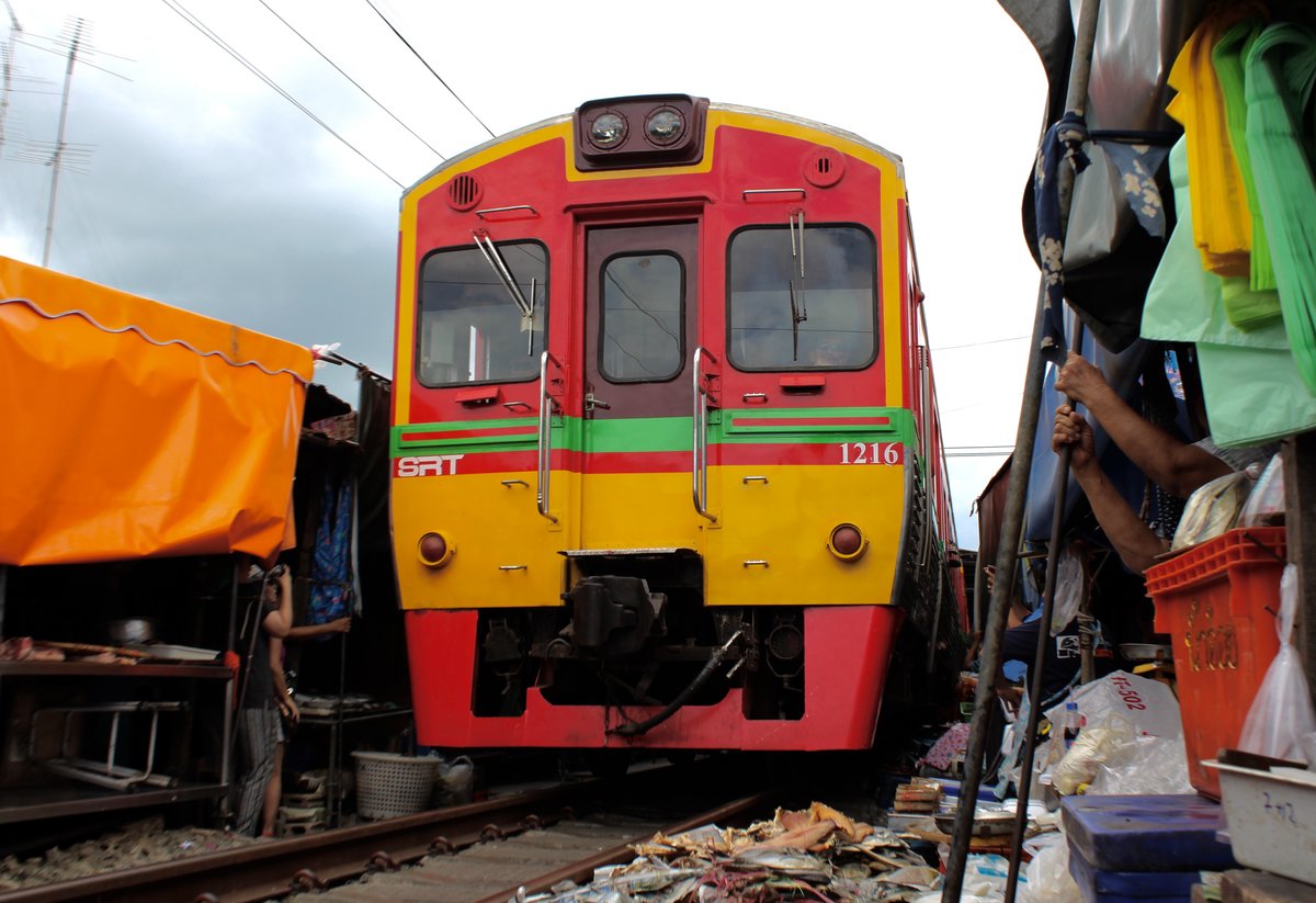 NKF 1216 bei seiner Fahrt als ORD 4384 durch den Markt beim Bf. Mae-Klong. Donnerstag, 3. August 2017

Der Mae Klong Markt stand seit über 100 Jahren an der genaugleichen Stelle, auch heute noch, als bevor hier eine Bahnstrecke hindurch gelegt wurde. Die Händler müssten dann eine Entscheidung treffen:  Soll der Markt
 umziehen oder bleiben , hatten sich Händler gefragt... Sie entschieden für's  bleiben  und  teilen  sich heute die Gleise mit der Maeklong Railway. Mittlerweile ist es wohl der gefährlichste Markt der Welt!